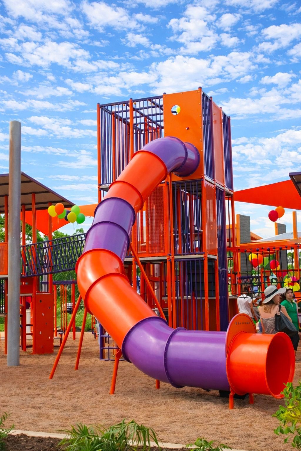 A vibrant orange and purple spiral tube slide stands in a sandy playground under a bright blue sky.