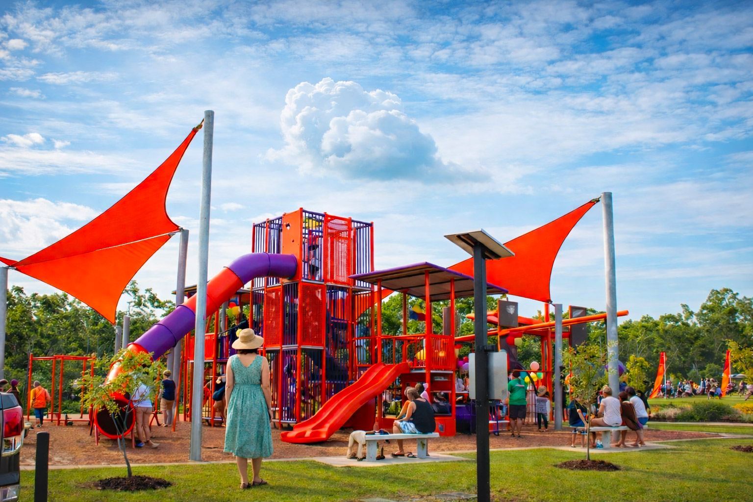A vibrant orange and purple playground structure with large shade sails, set in a sunny park with people nearby.