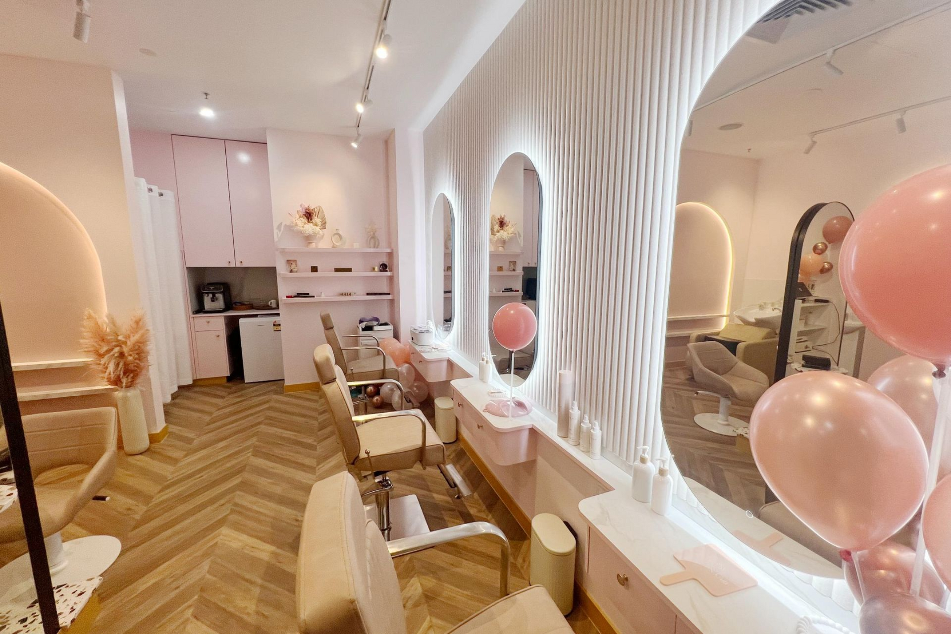 A chic salon interior featuring a central double-sided mirror, beige swivel chairs, a curved bench, and soft pink decor.