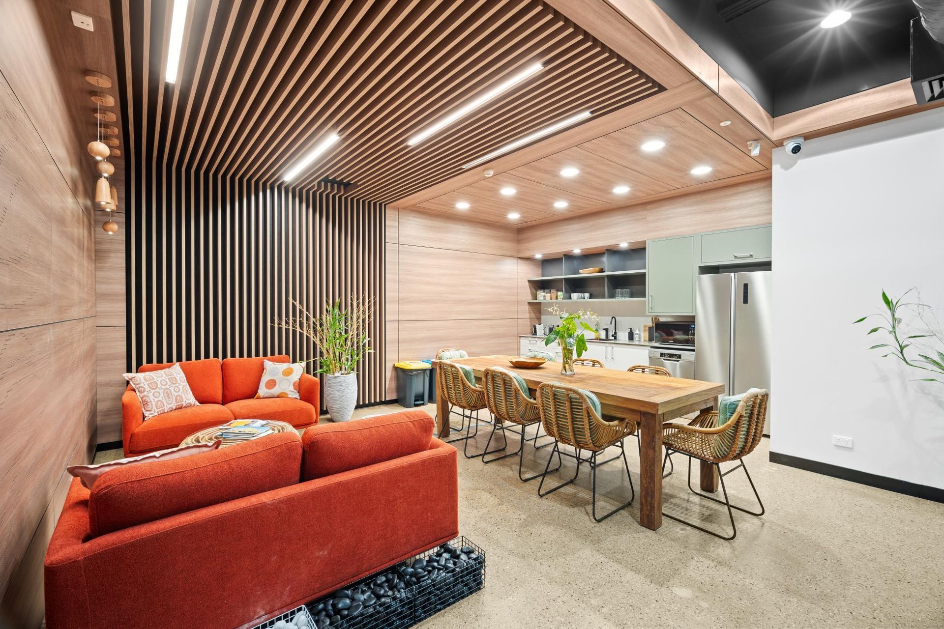Modern lounge and kitchen area with orange sofas, a wooden dining table, slatted wood ceiling, and recessed lighting.