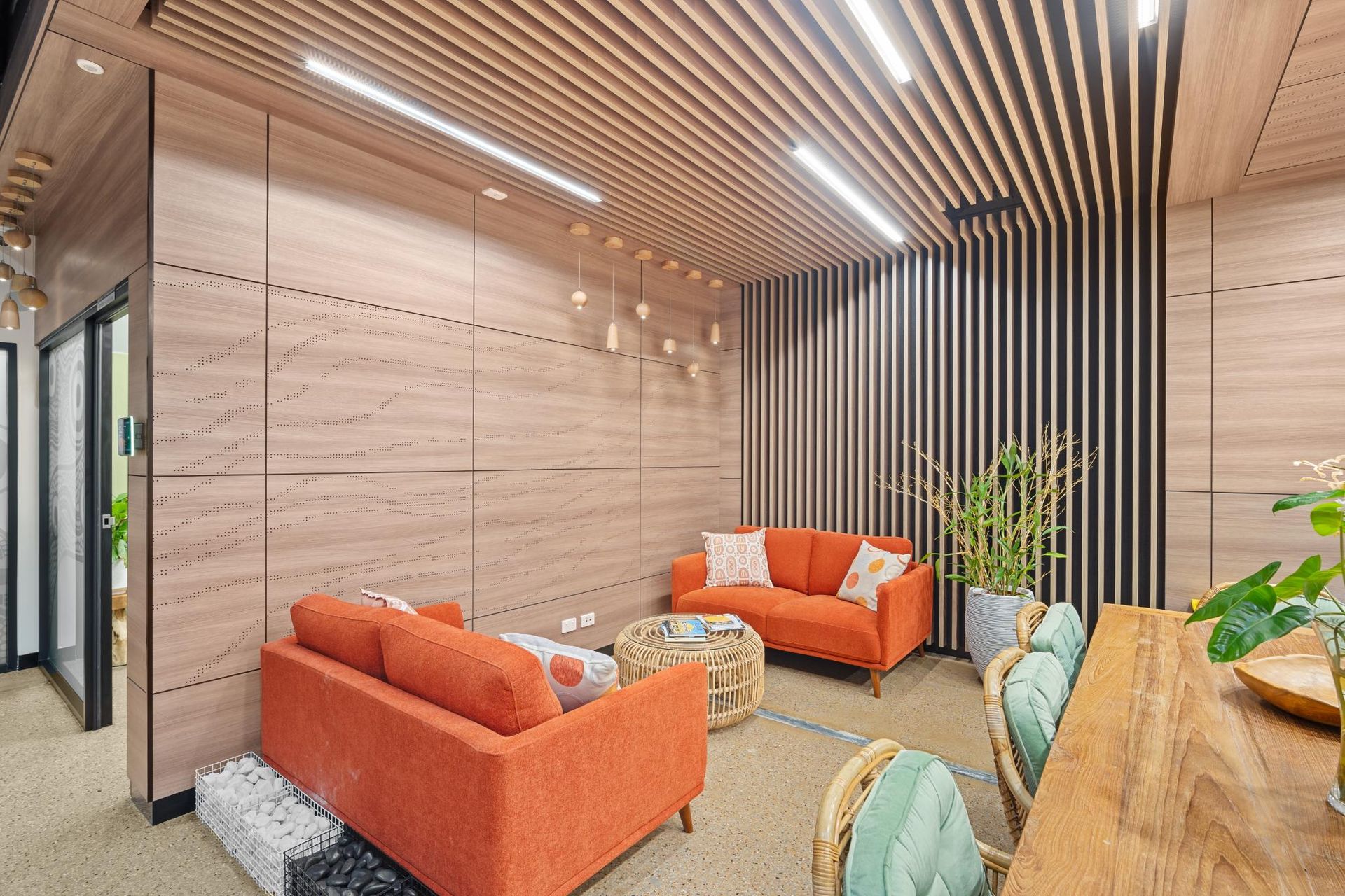 Modern office lounge with light wood-paneled walls, orange sofas, and a slatted wood ceiling with LED strip lighting.