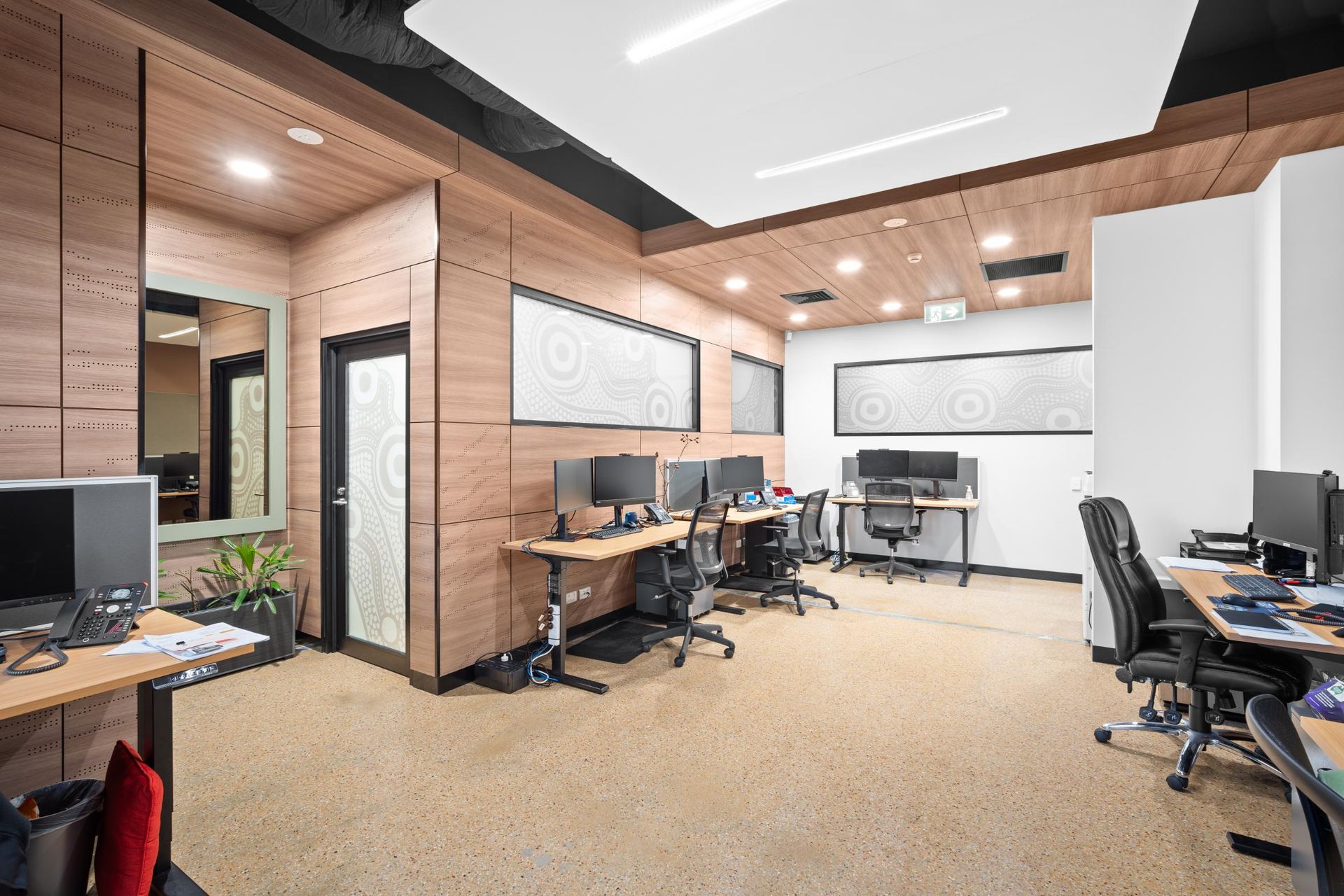 Modern office space with multiple workstation desks, ergonomic chairs, wood-paneled walls, and recessed ceiling lights.