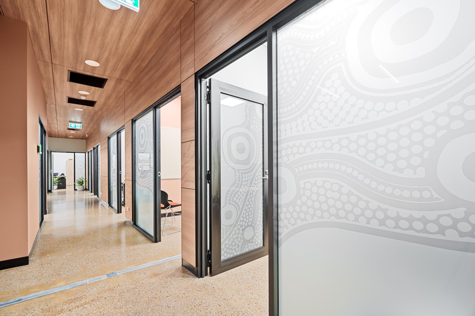 A modern office hallway with wood-paneled ceilings, terrazzo floors, and frosted glass doors featuring abstract art.