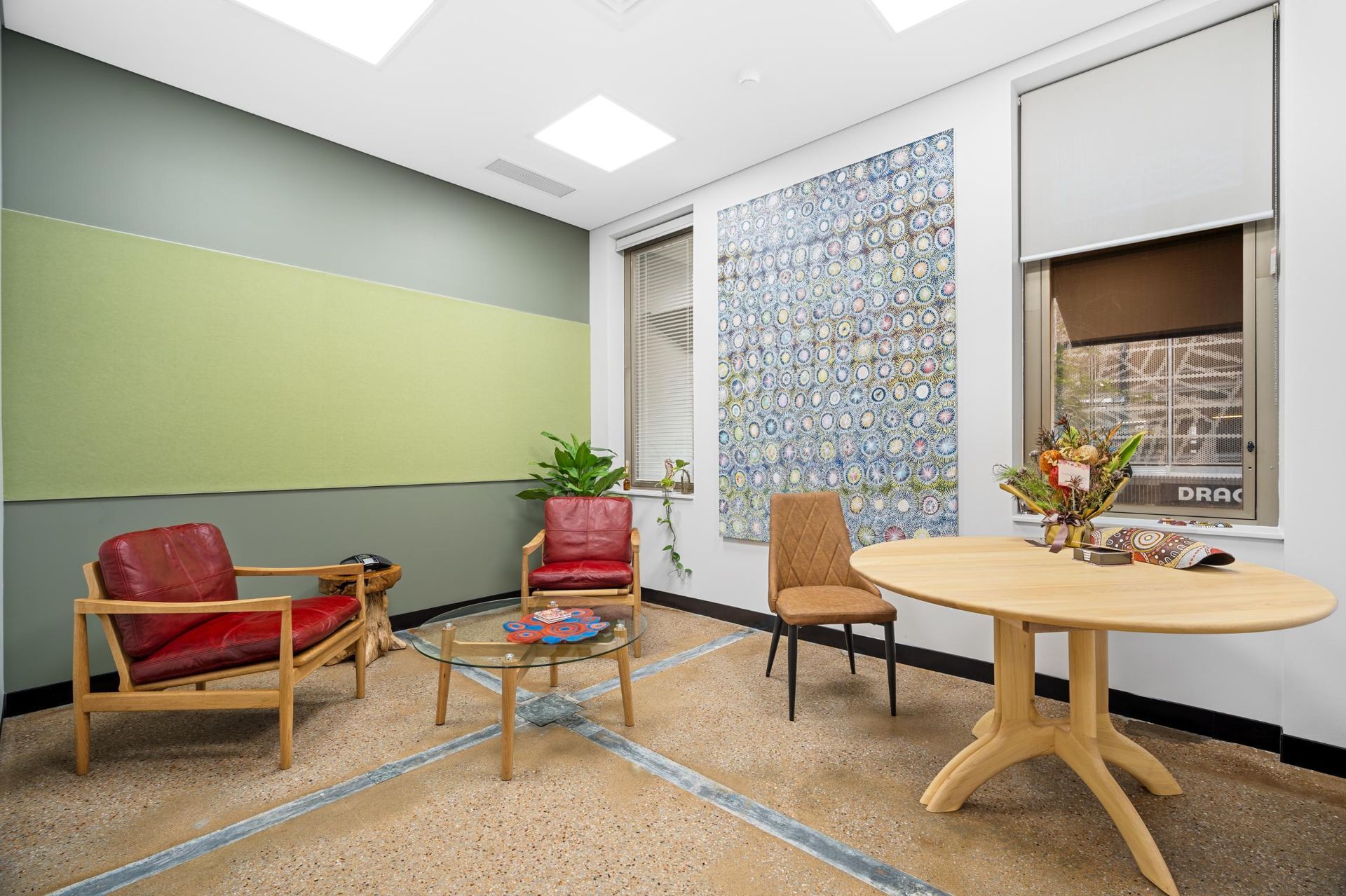 A modern office lounge with two red armchairs, a glass coffee table, a wood table, and a brown chair on speckled flooring.