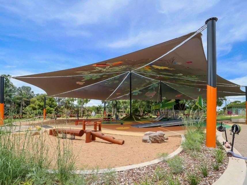 A shaded playground with a large brown canopy, wooden balance logs, and sandy ground under a bright blue sky.