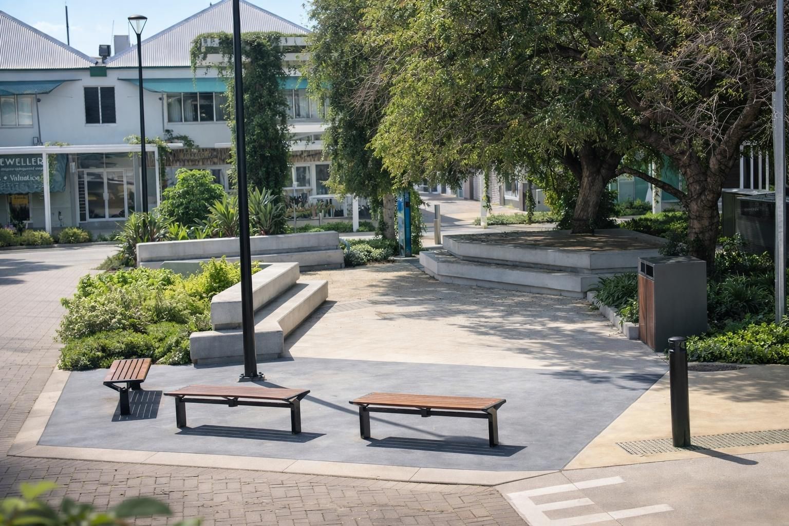 A sunny, paved outdoor plaza featuring wooden benches, concrete tiered seating, greenery, and a large tree.