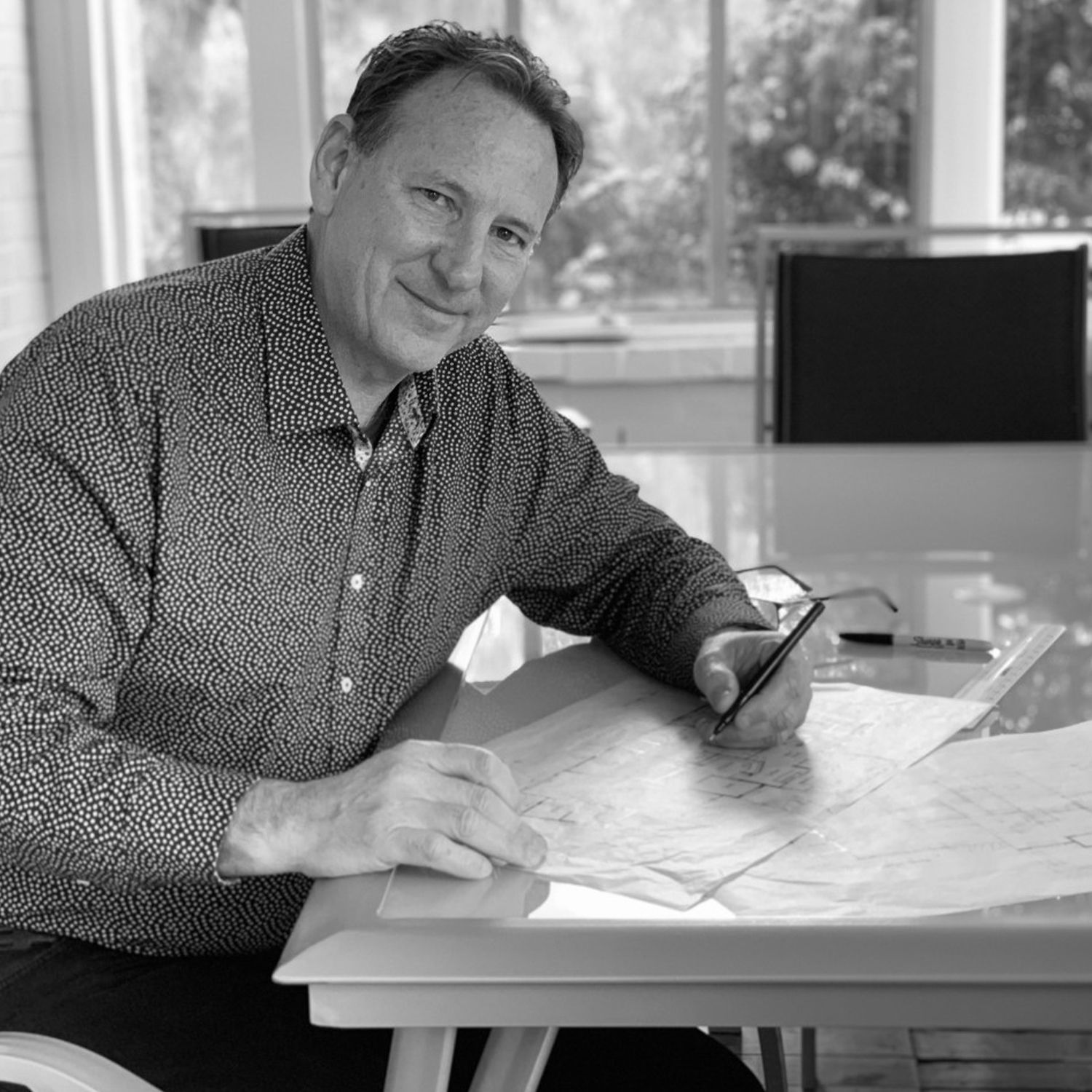 A person in a patterned button-down shirt smiles while working on sketches at a desk near a window.