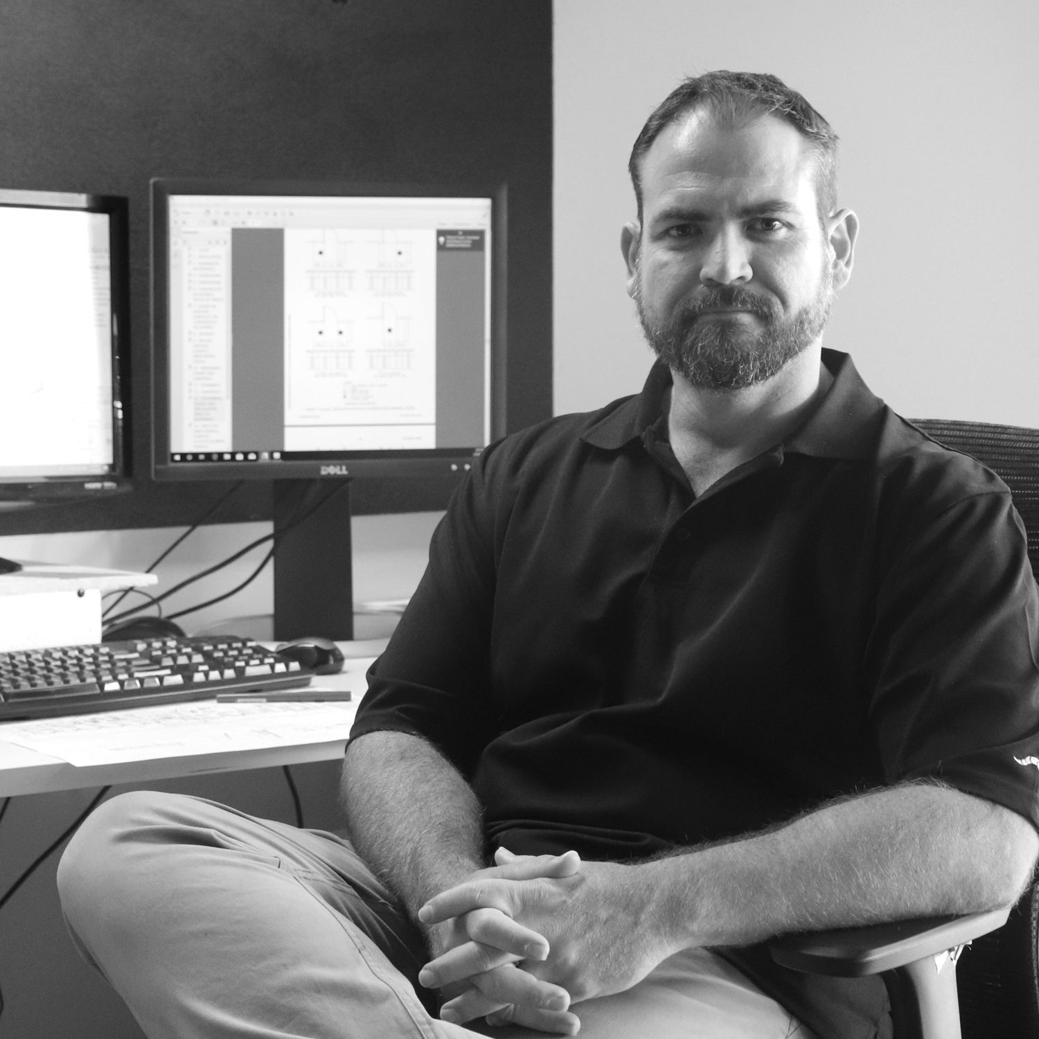 A bearded person sitting at a desk in an office with two computer monitors, hands clasped, looking at the camera.
