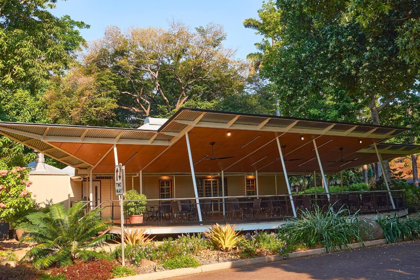A modern, open-air pavilion with a sweeping, angular wooden roof, surrounded by lush green trees and garden landscaping.