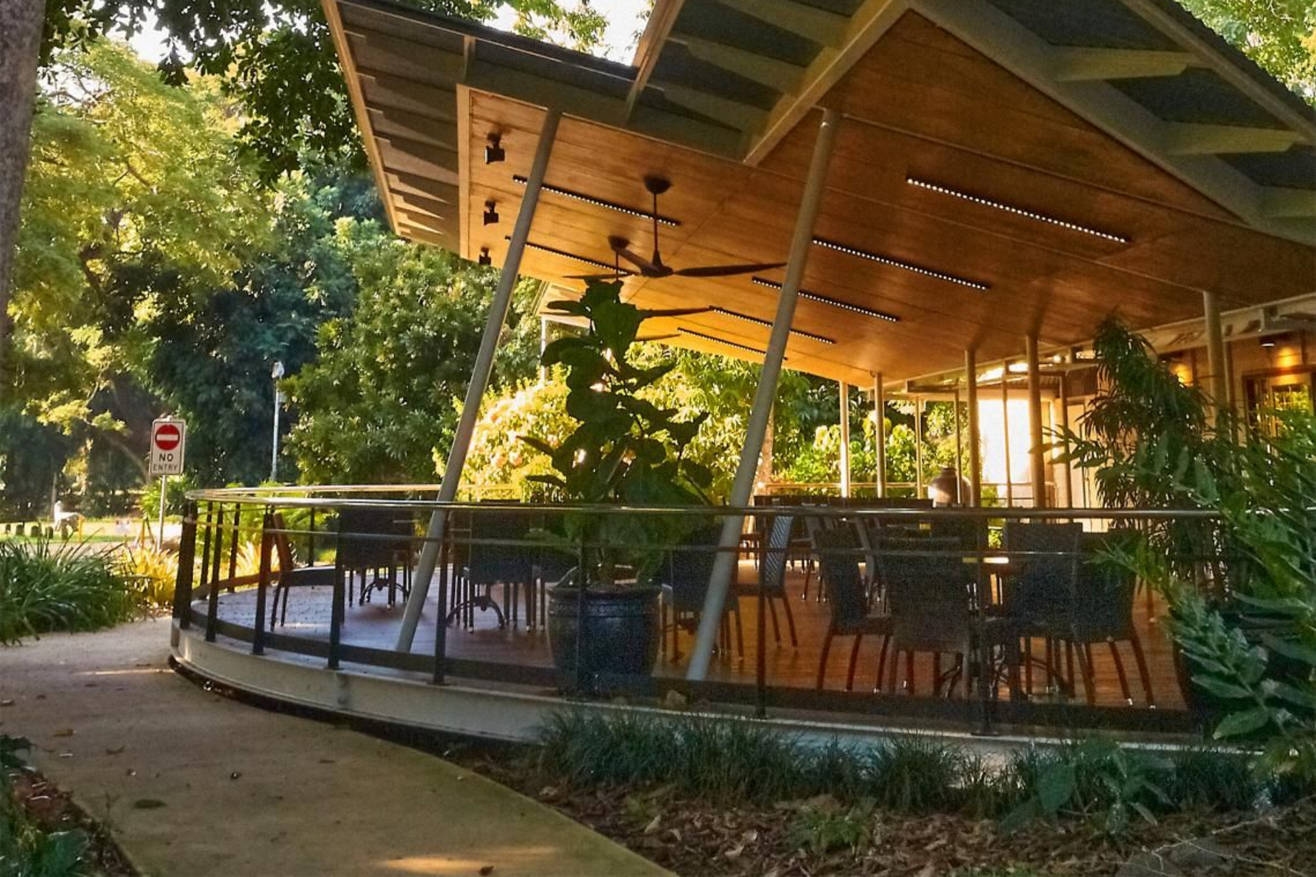 A modern, open-air pavilion with a sweeping, angular wooden roof, surrounded by lush green trees and garden landscaping.
