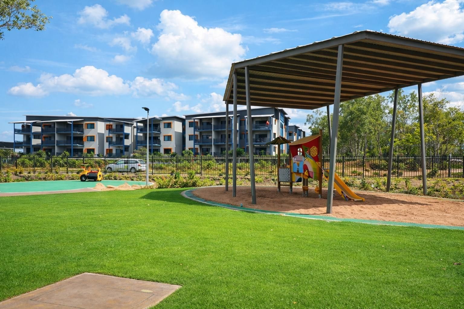 A playground with a covered slide structure on a sandy patch, situated next to a green lawn and apartment buildings.