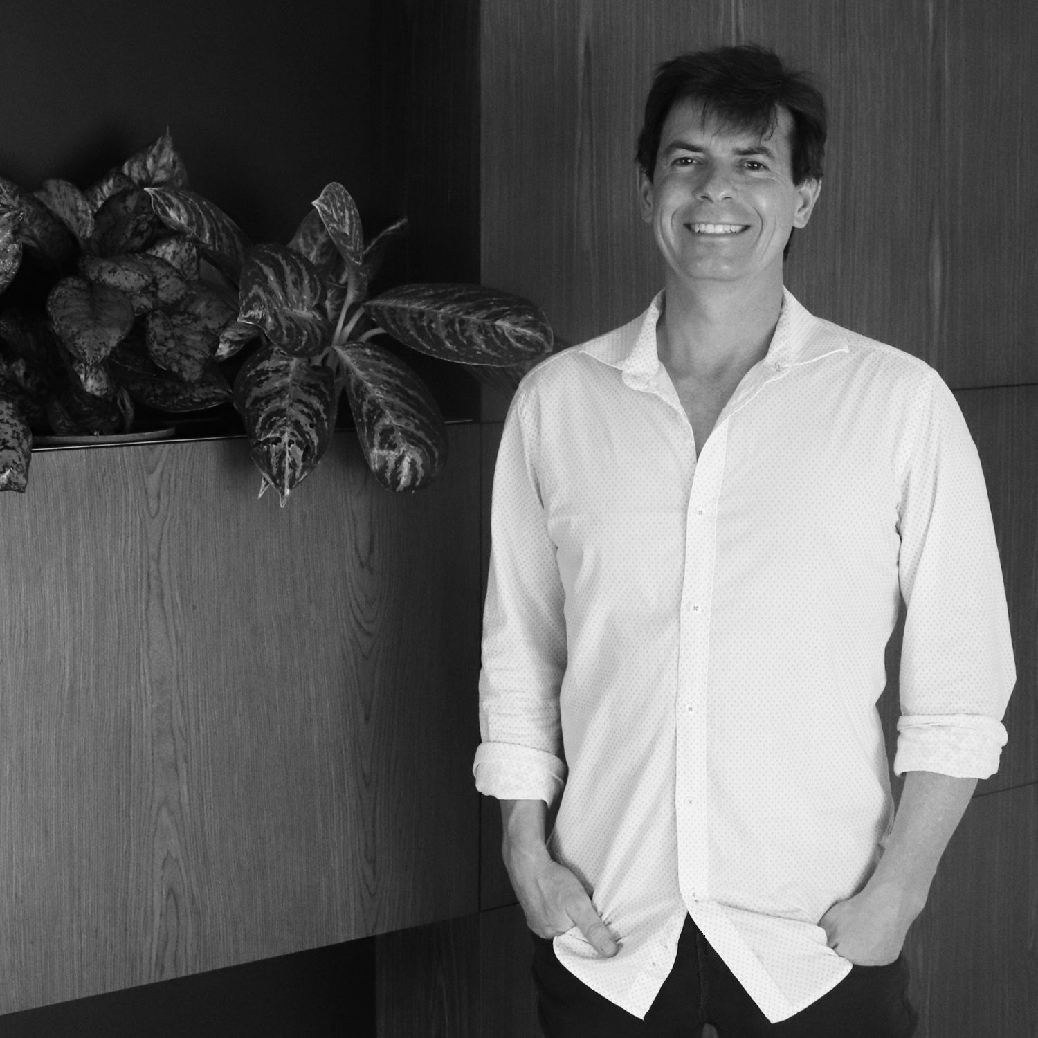 A smiling person stands with hands in their pockets next to a large potted plant against a dark wood paneled wall.