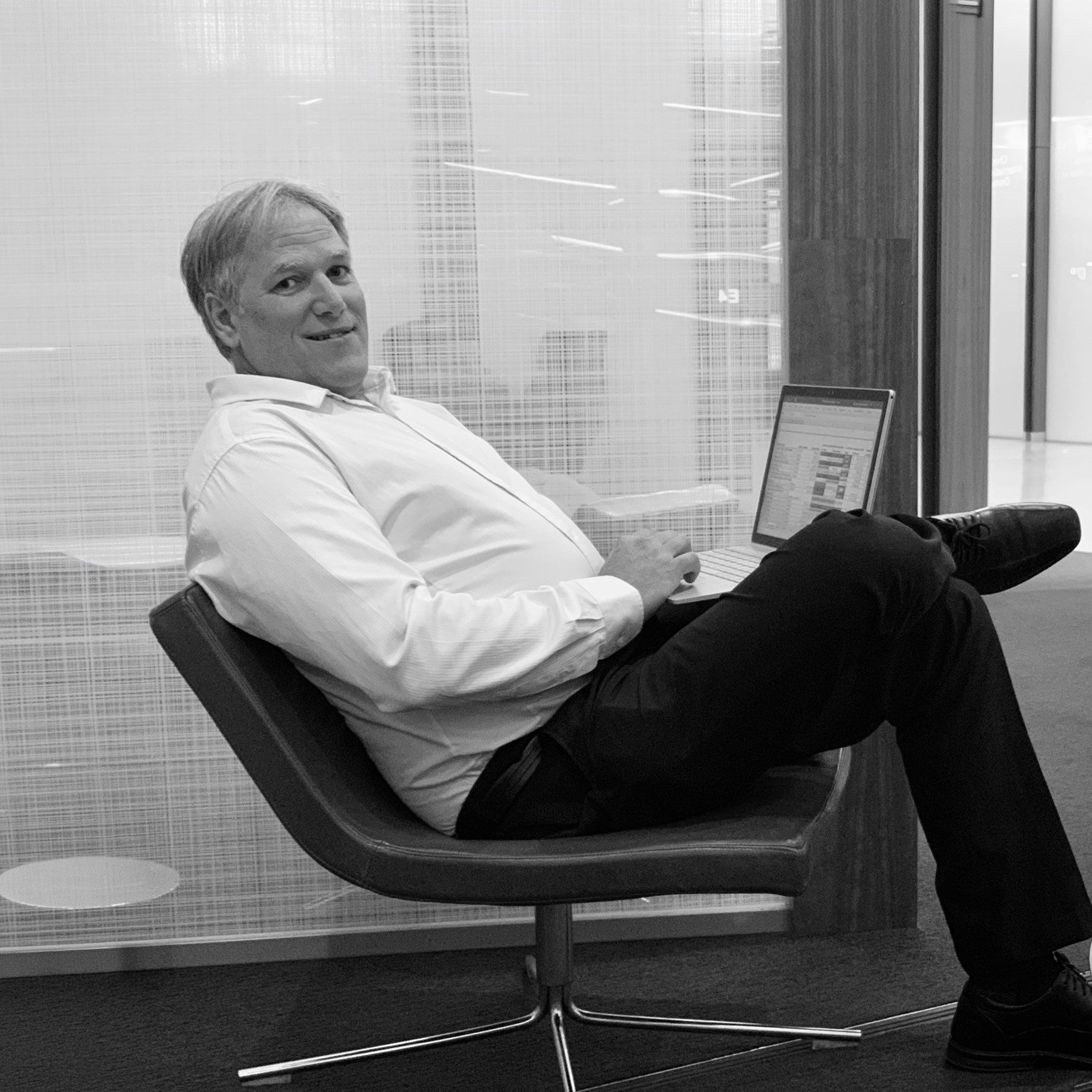 A person in a white button-down shirt and dark pants sits in a modern chair working on a laptop in a bright office.