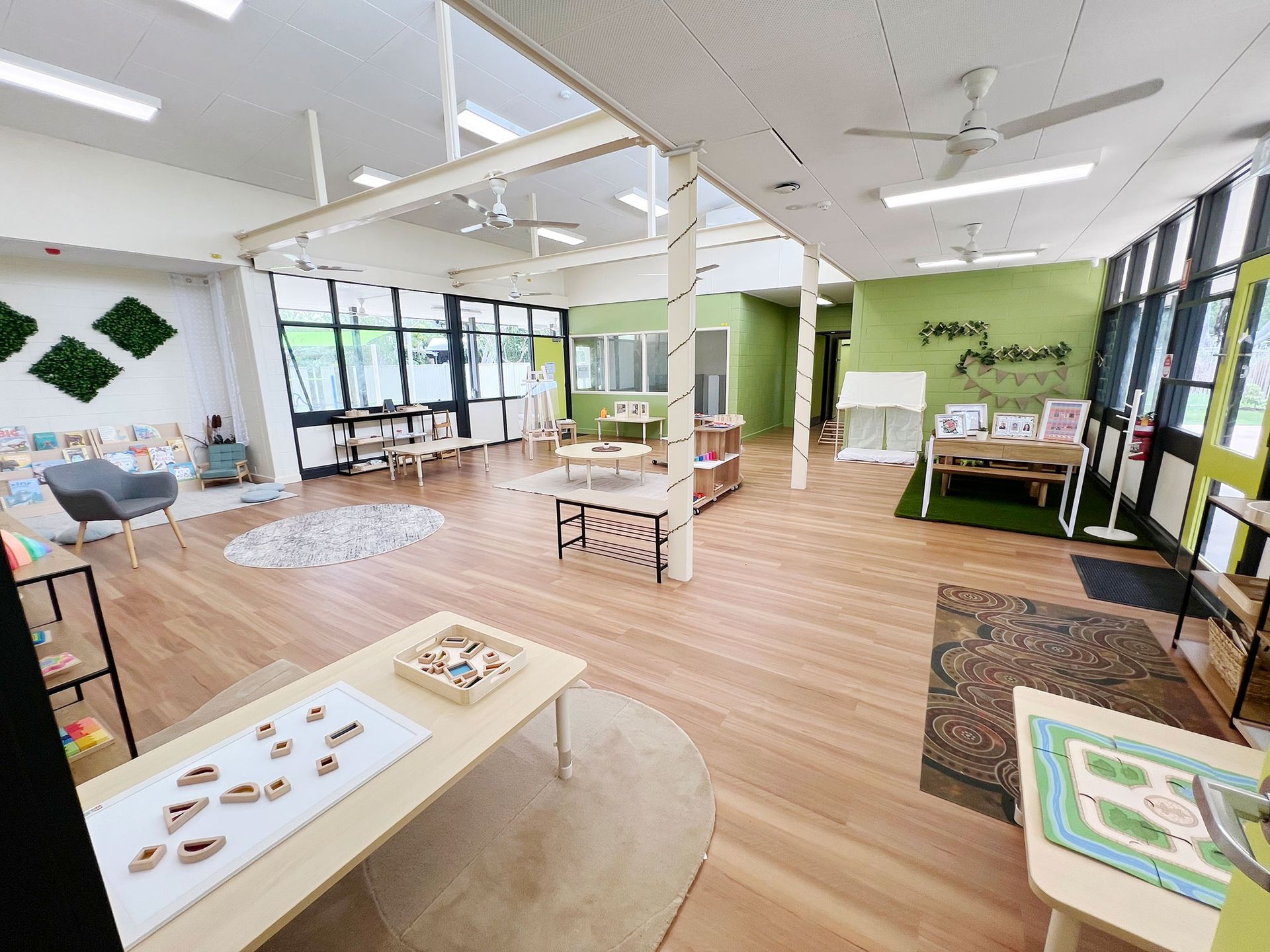 A bright, modern childcare space with wooden floors, low activity tables, natural light, and green accent walls.
