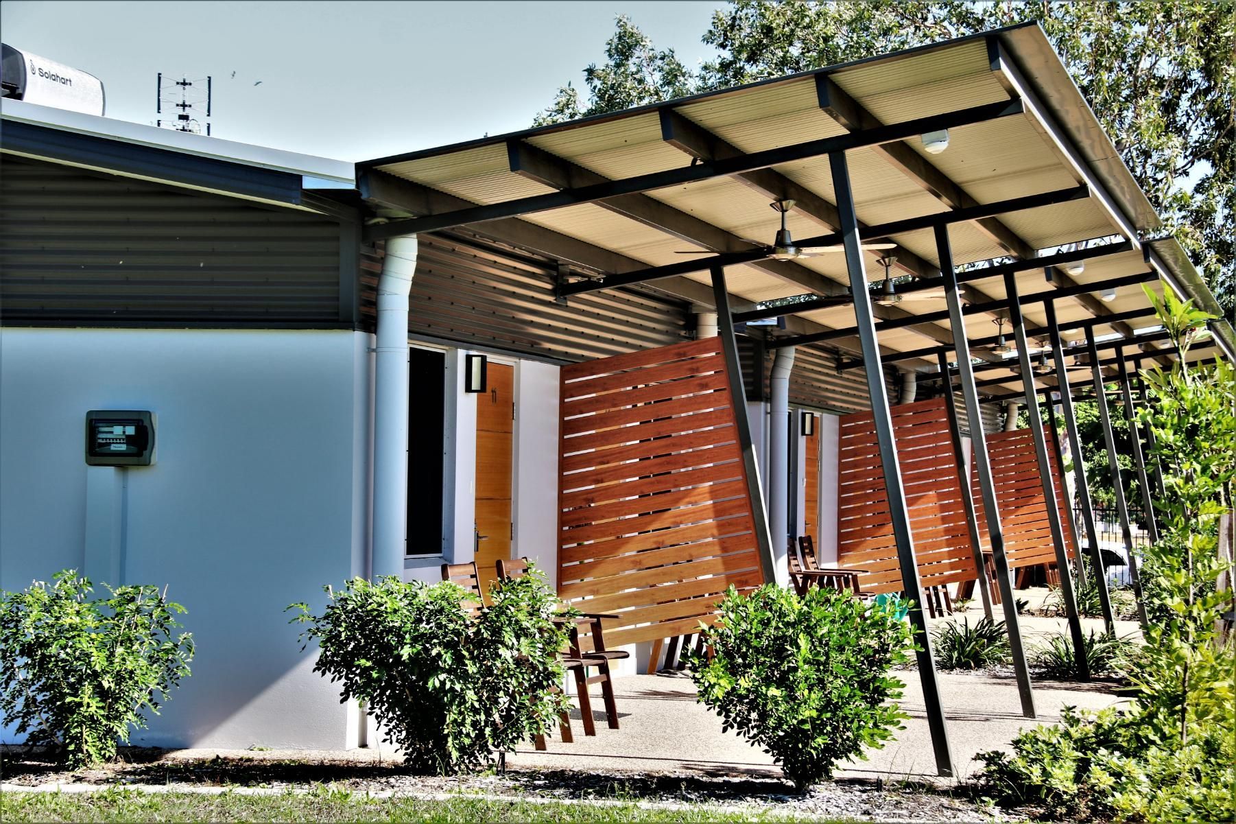 A white building with wooden privacy screens under a corrugated metal awning, surrounded by green shrubs.