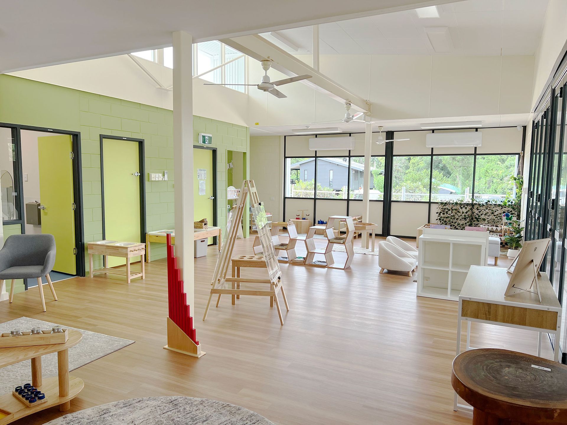 A spacious, brightly lit preschool classroom with light-colored furniture, wooden flooring, and soft green accent walls.