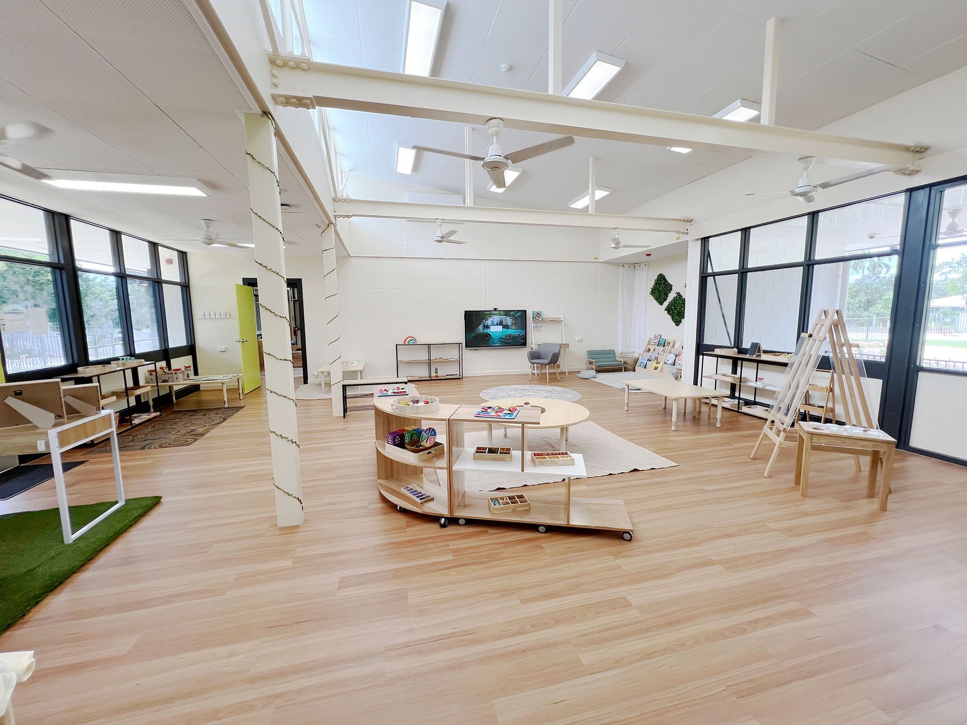 A brightly lit, modern open-plan preschool or classroom with wooden floors, low shelves, a television, and large windows.