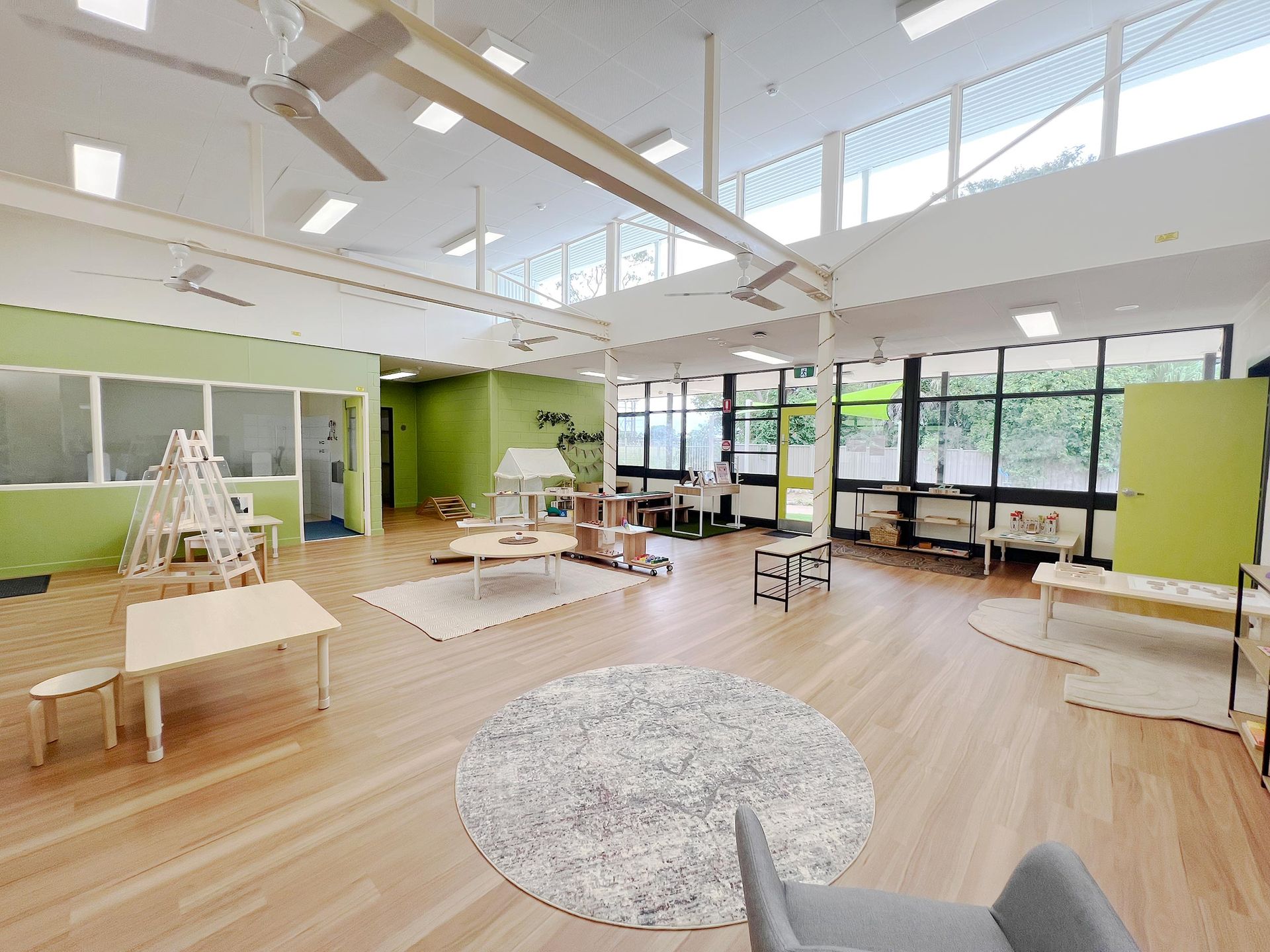 A spacious, sunlit preschool classroom with light wood floors, green walls, wooden furniture, and circular area rugs.
