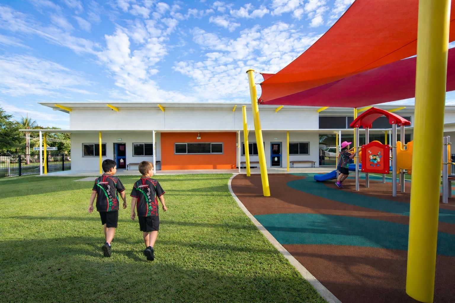 Two children walk across a grassy school playground toward a modern building with a colorful shade structure.