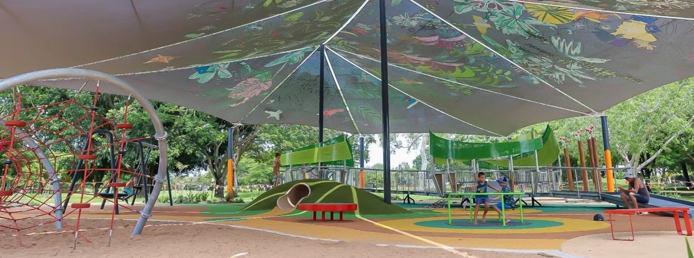 Shaded playground with colorful play structures, slides, swings, and trees under a large canopy.
