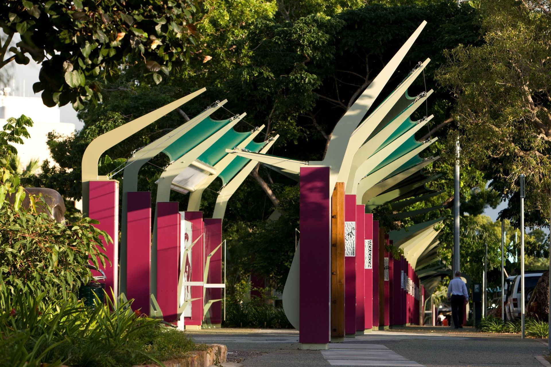 Outdoor public walkway featuring modern, magenta pillars supporting arched, green-and-white angled canopies in a park.