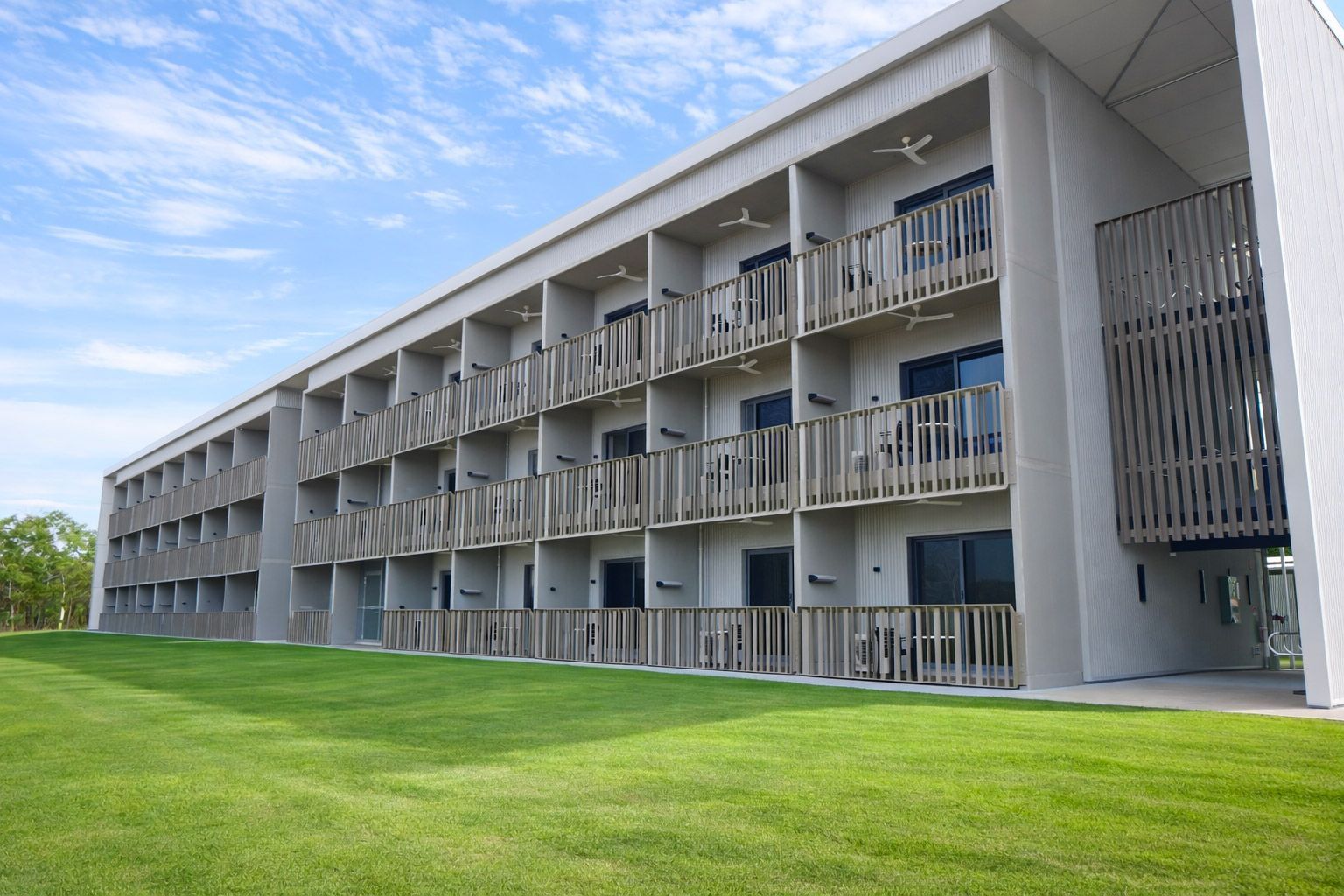 A three-story modern building with gray walls, wooden balcony railings, and dark glass doors, set against a lawn and sky.