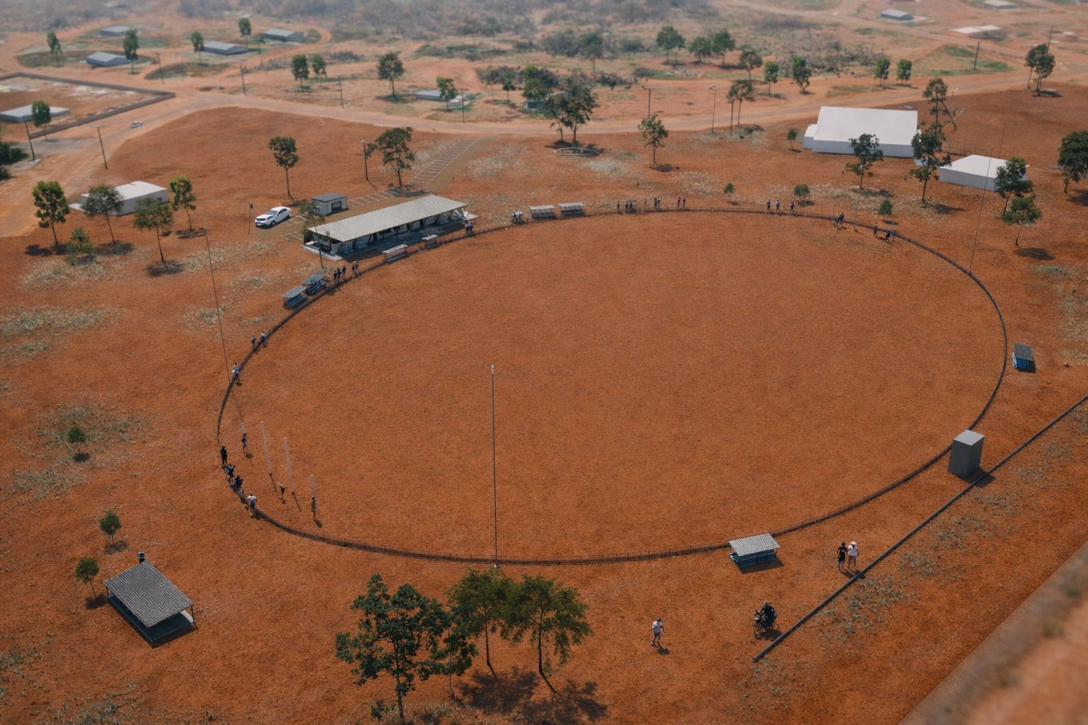 High-angle shot of people standing along a large, circular dirt track in a dry, open field with sparse trees and buildings.