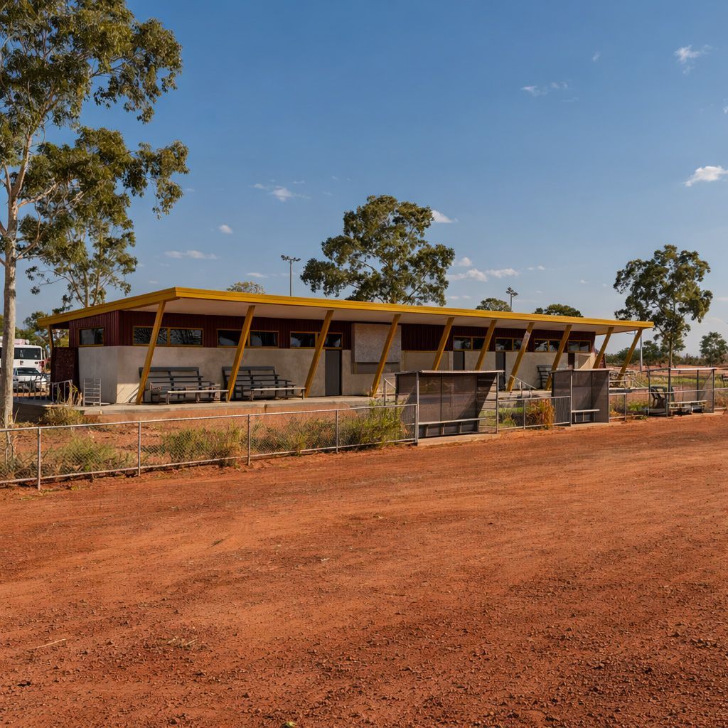 A modern, single-story community building with a yellow roof and red walls stands next to a red dirt field under blue sky.