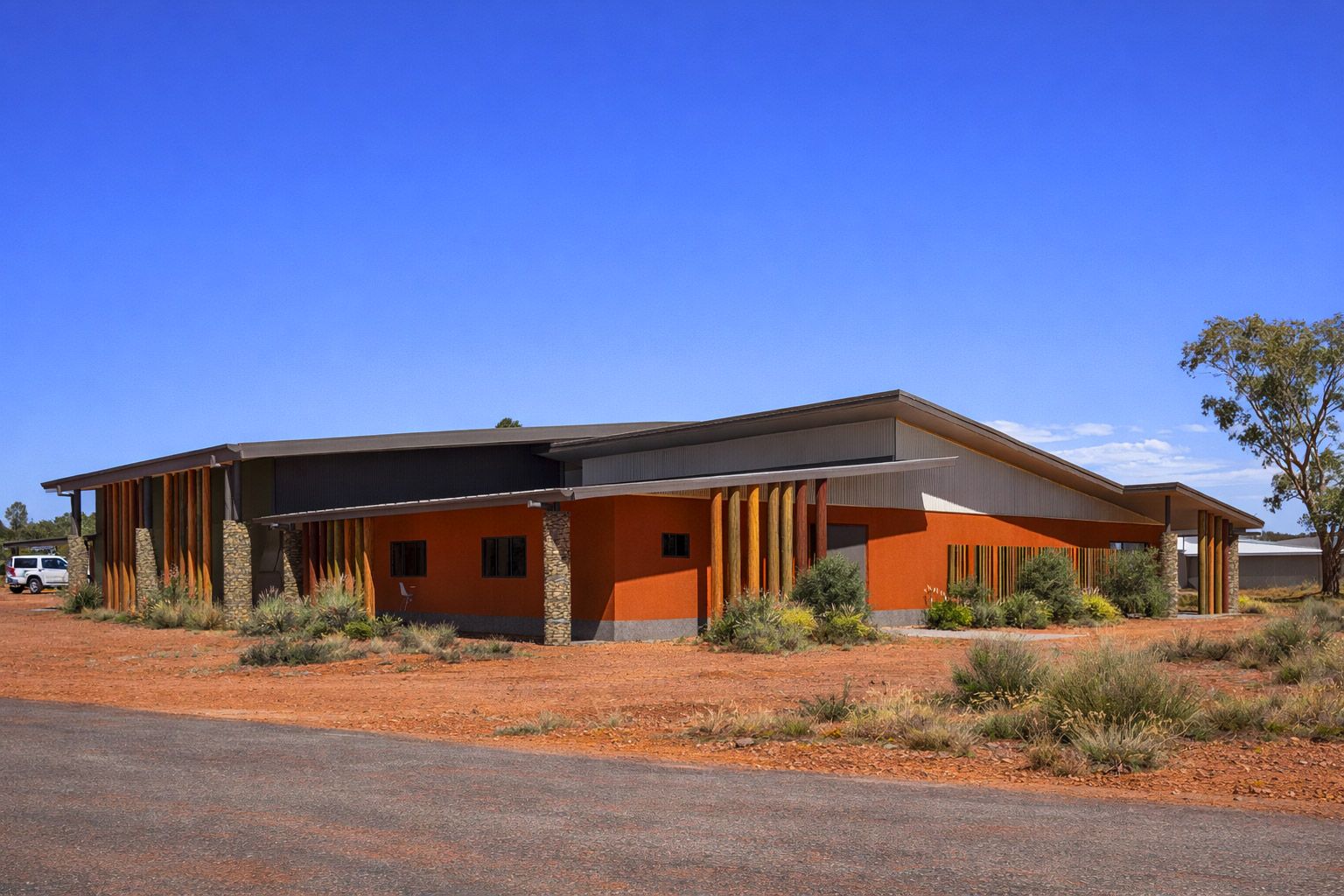 A low, modern building with orange walls and wooden pillars stands in a dry, red-earthed landscape under a clear blue sky.