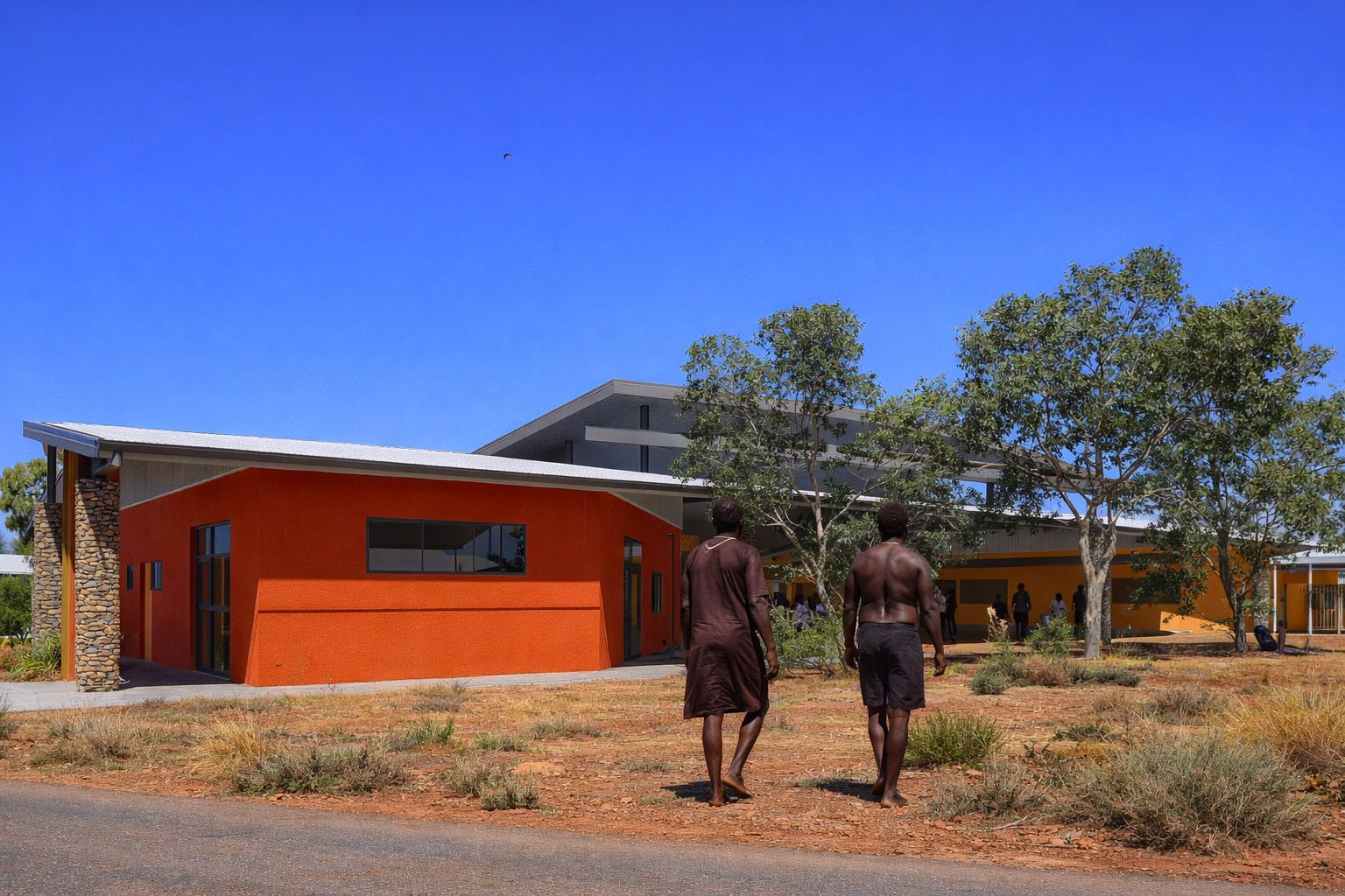 Two people walk across a dry, brushy landscape toward a modern building with a bright orange exterior wall.