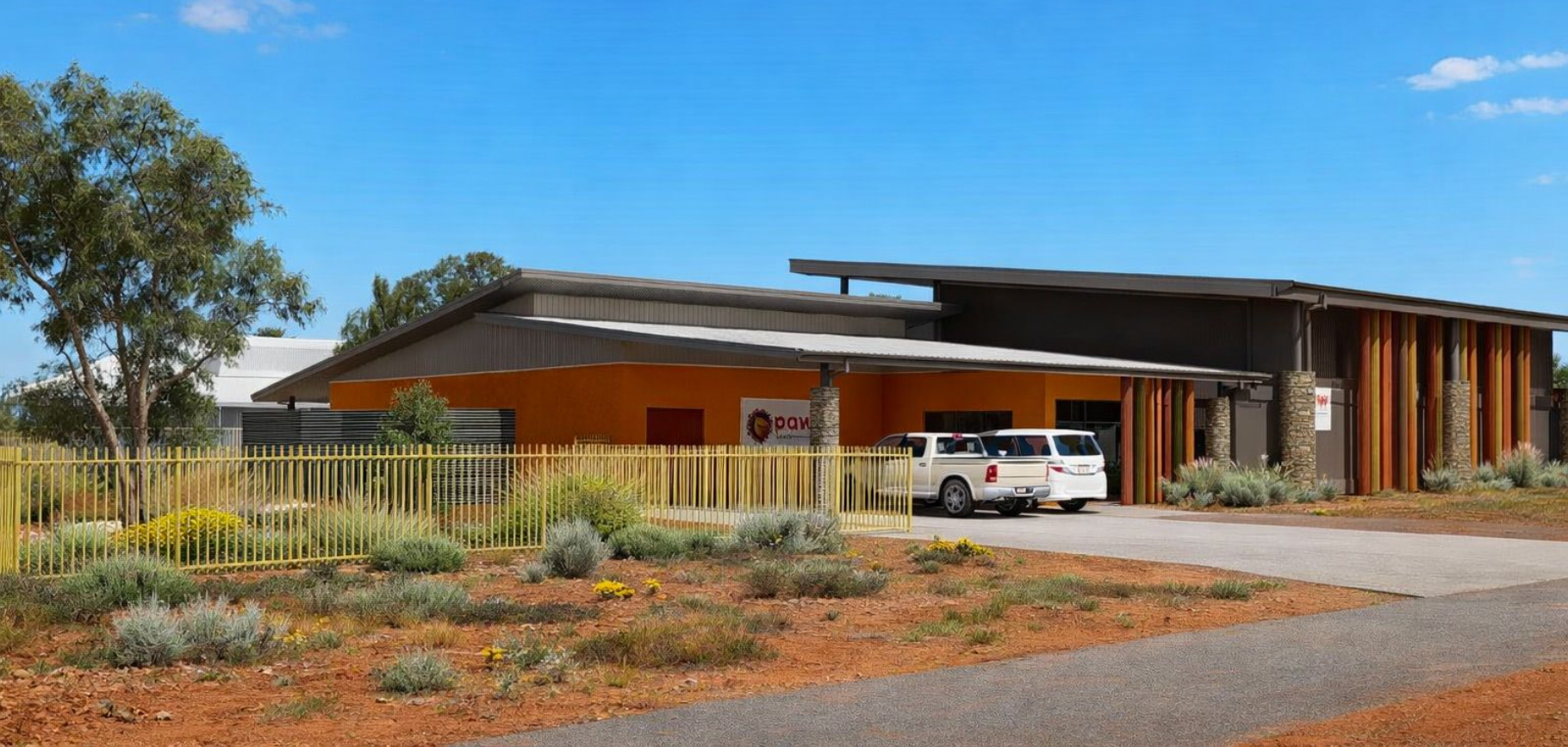 A low-slung, modern building with orange walls and wood accents under a clear blue sky, surrounded by arid landscaping.