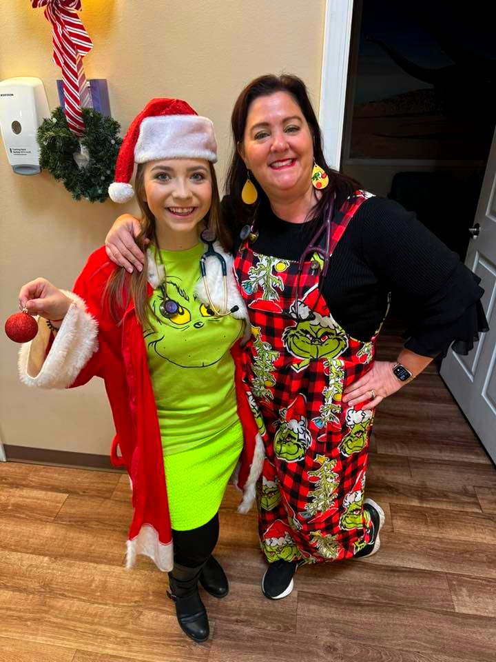 Two women in festive holiday outfits smiling indoors, one in a Santa hat, arm in arm.