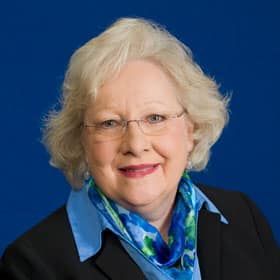 A woman wearing glasses and a blue scarf is standing in front of a blue background.