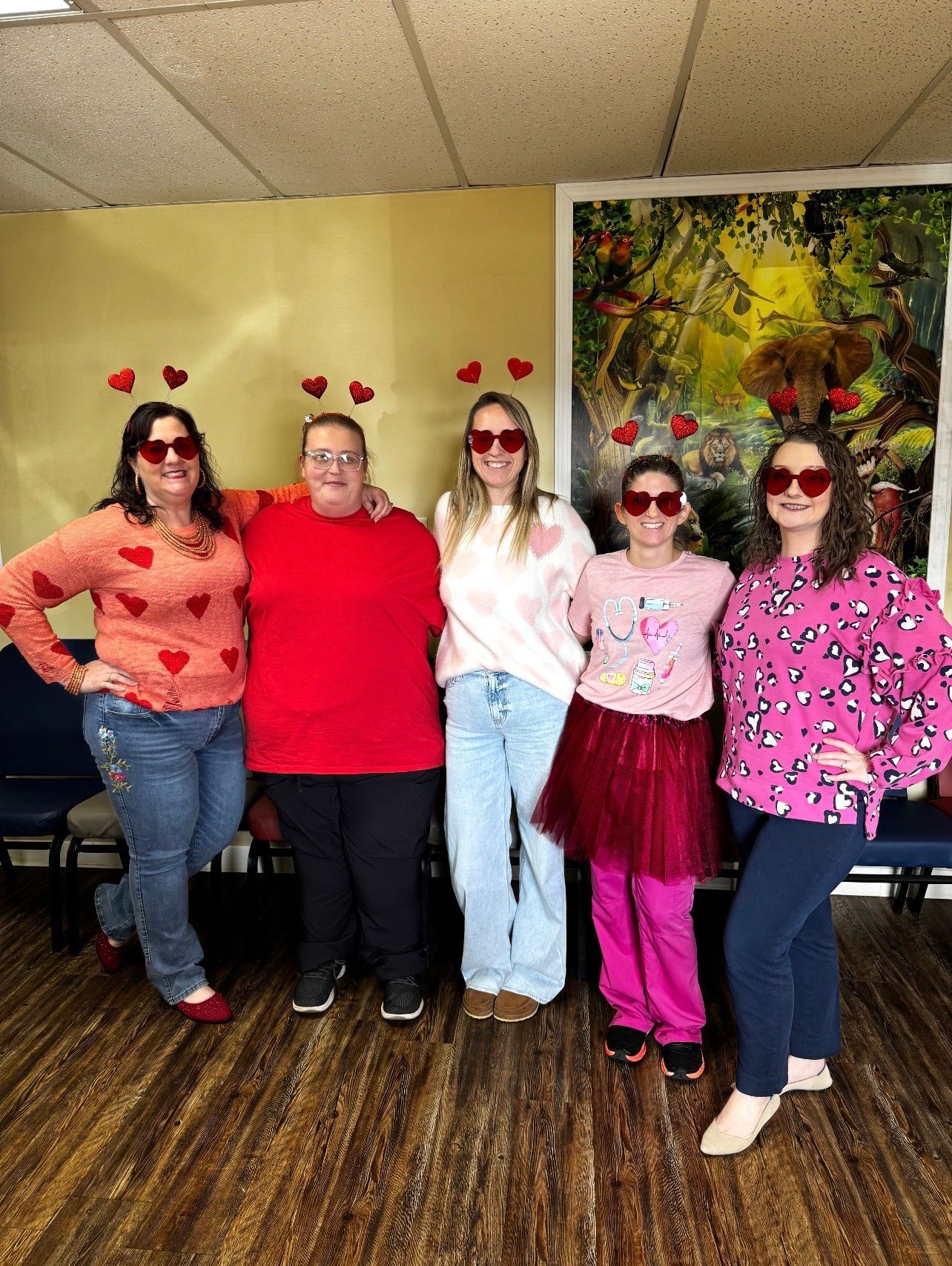 Five people posing indoors, four in red-themed outfits with heart headbands, in front of a colorful mural.