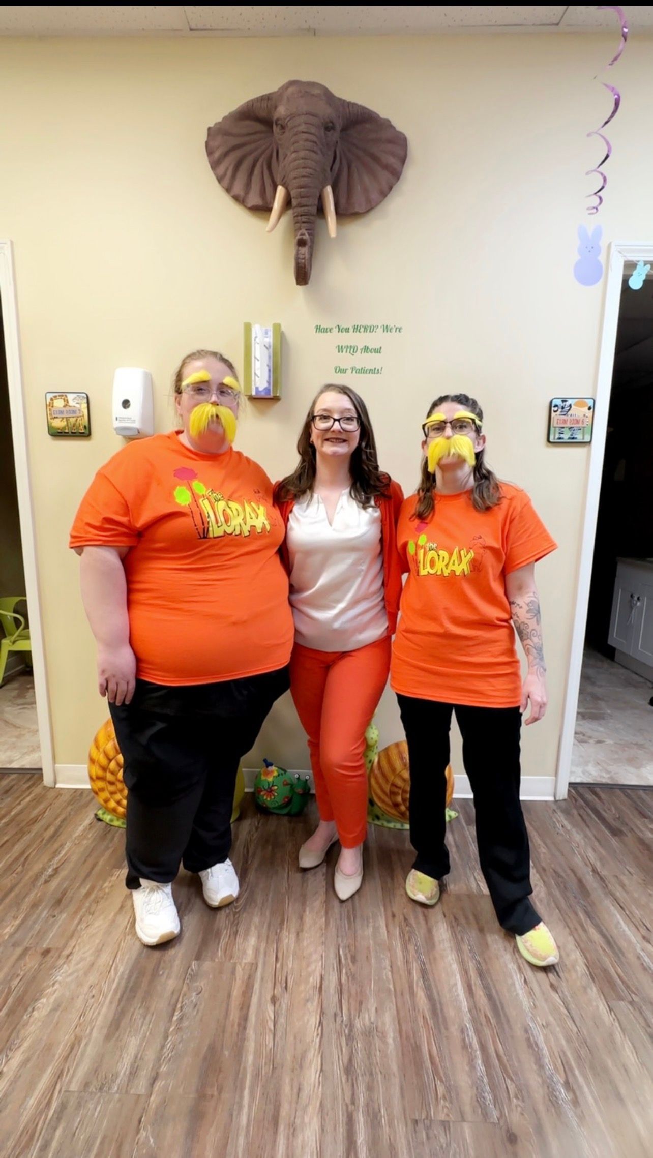 Three people posing indoors beneath a mounted elephant head, with balloons and wall decorations behind them.