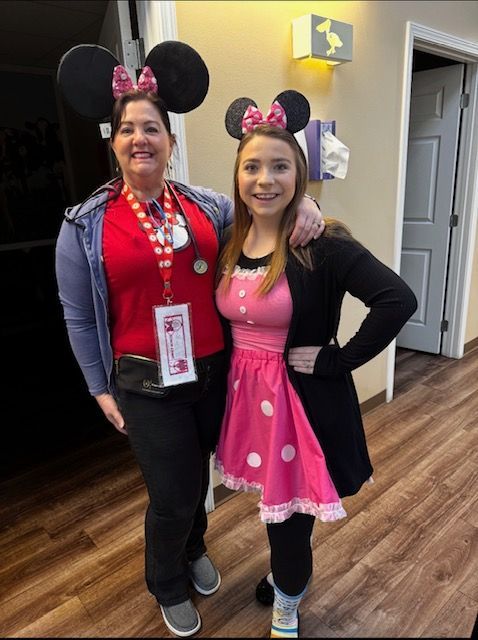 Two women in pink Minnie Mouse outfits stand smiling in a hallway.