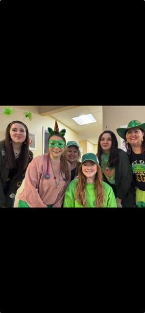 Five smiling people in green St. Patrick’s Day hats and shirts pose indoors with shamrock decorations.