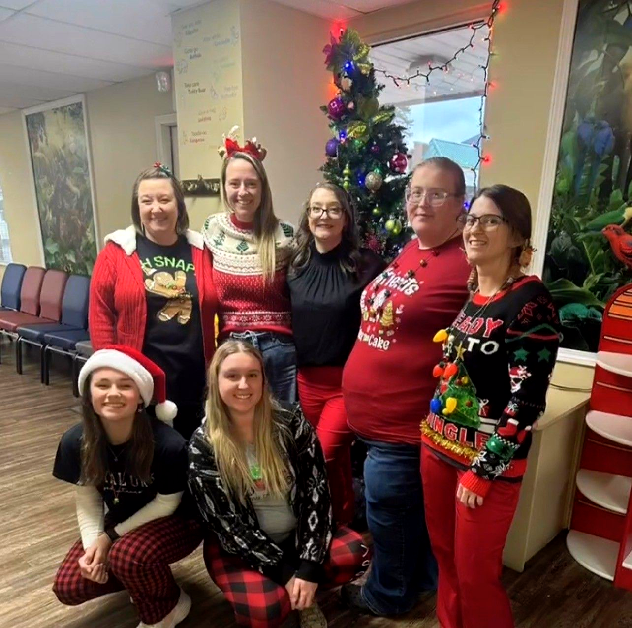Seven people pose indoors by a decorated Christmas tree in festive holiday sweaters.