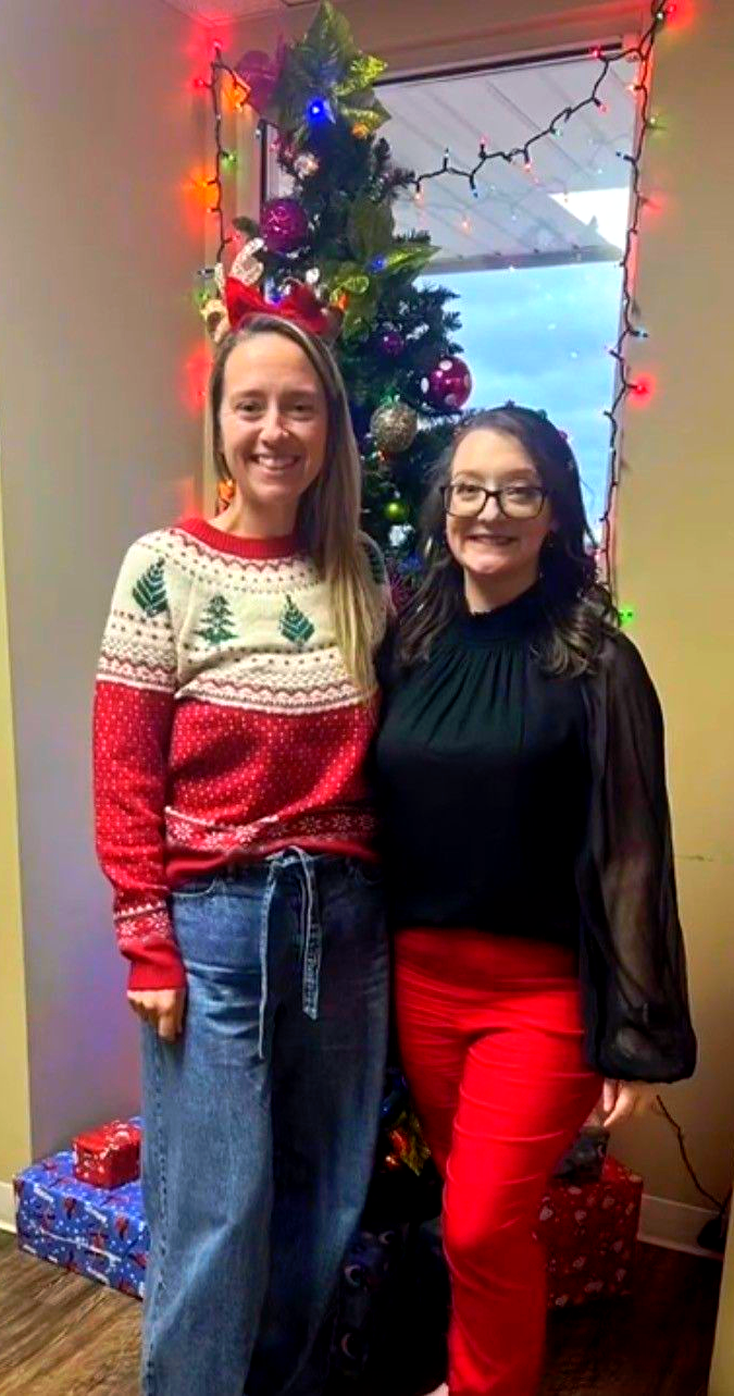 Two women standing beside a decorated Christmas tree, smiling in festive red and black outfits.