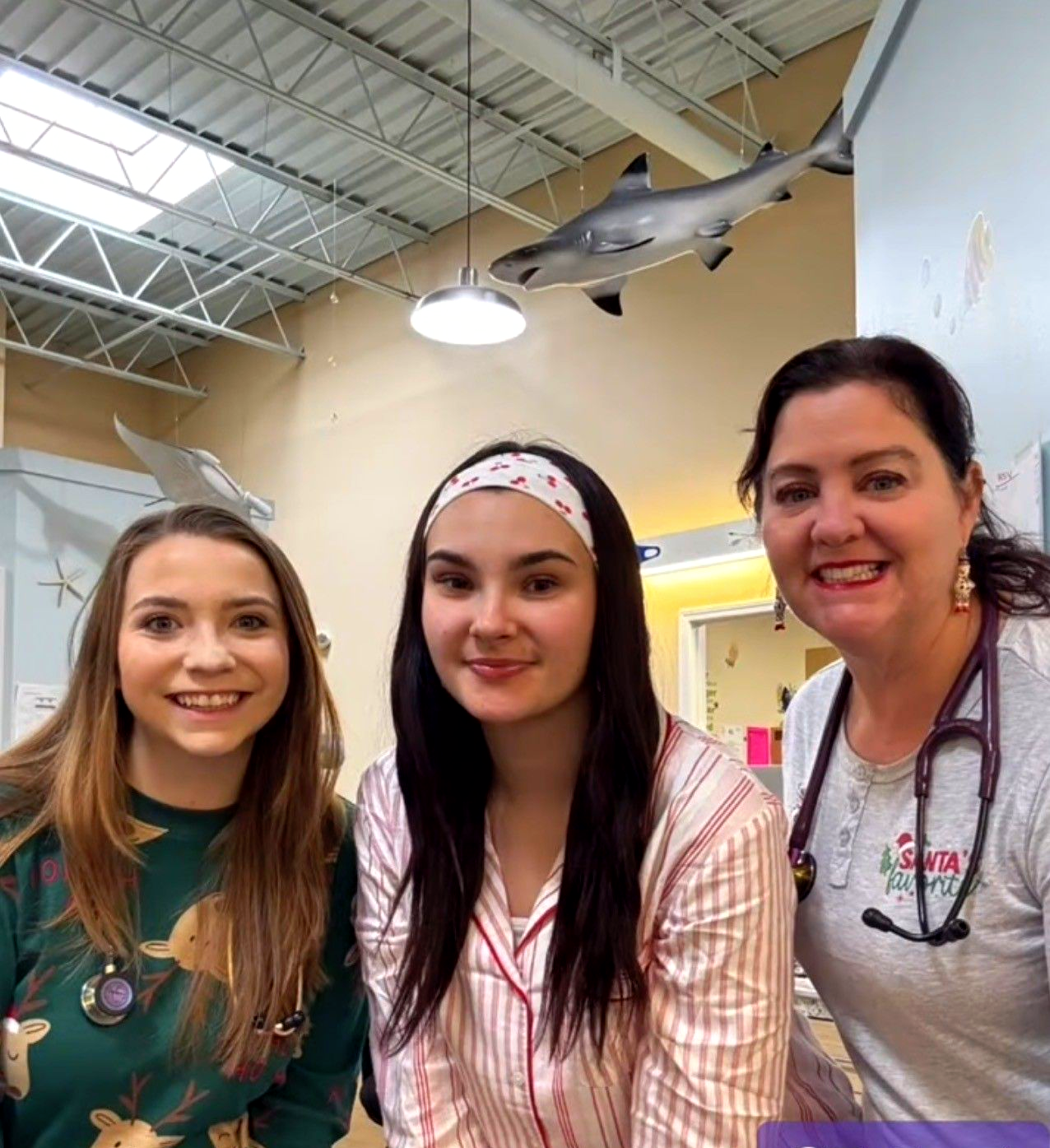 Three people smiling in a bright indoor space with a shark model hanging overhead.