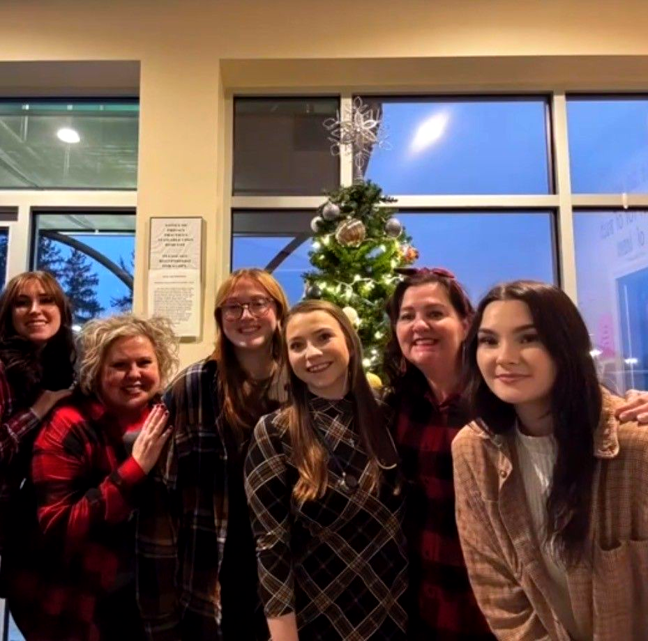 Six people posing indoors beside a decorated Christmas tree and large windows