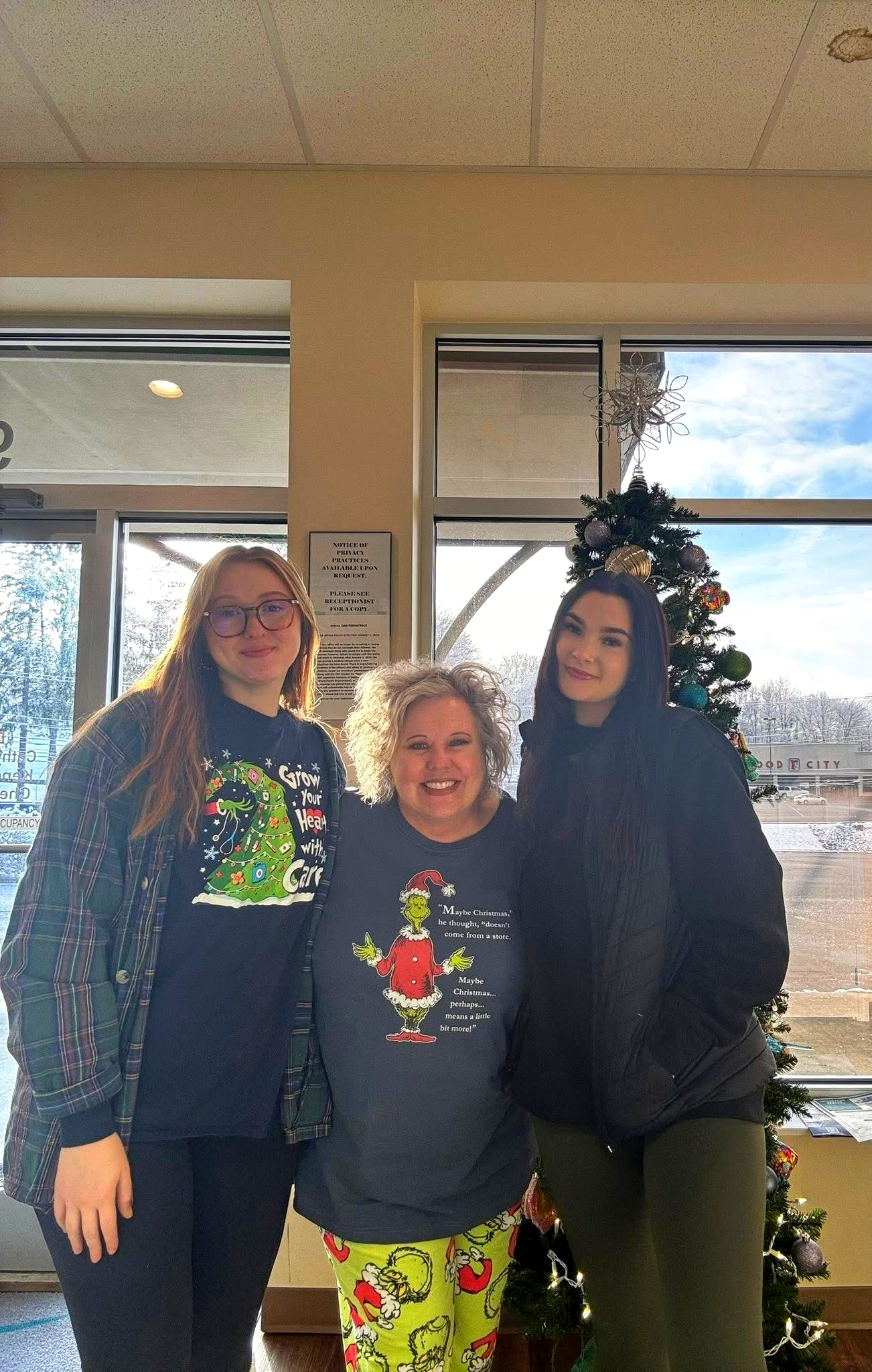 Three people standing indoors by a large window, smiling in front of a Christmas tree.