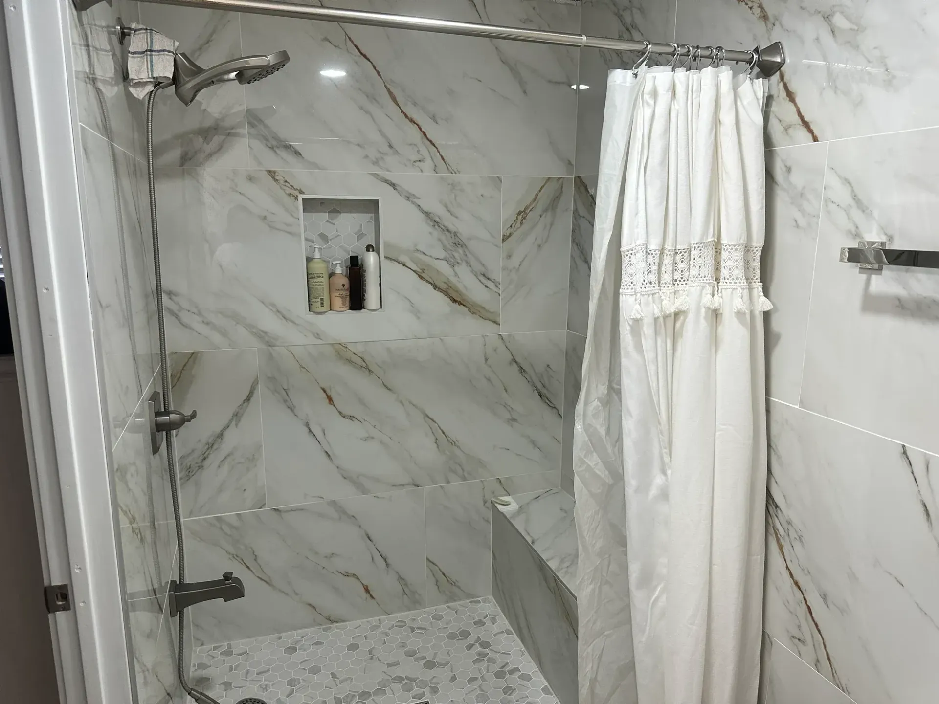 Modern marble-tiled shower with a white curtain, showerhead, and built-in shelf with toiletries