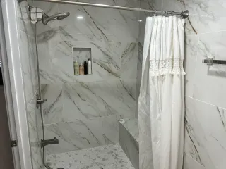 Modern marble-tiled shower with a white curtain, showerhead, and built-in shelf with toiletries