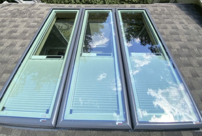 Three open blue-framed skylight windows on a shingled roof, reflecting clouds and sky