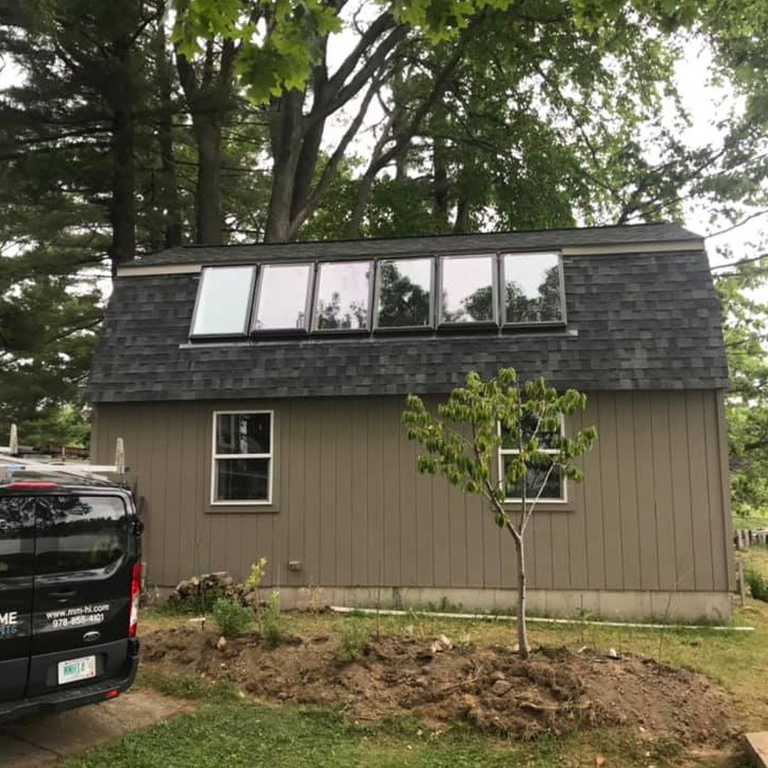 Small gray shed-style house with dark roof and dormer windows beside a parked black van, surrounded by trees.