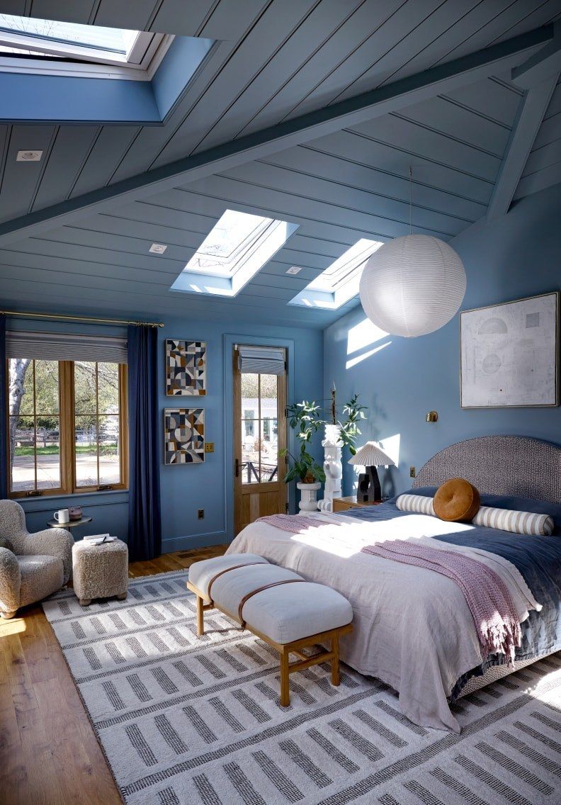 Blue bedroom with skylights, a bed, patterned rug, woven chair, and large windows overlooking greenery