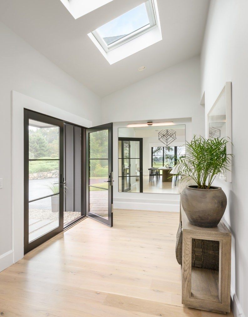 Bright modern entryway with open glass doors, skylight, light wood floors, and potted plants