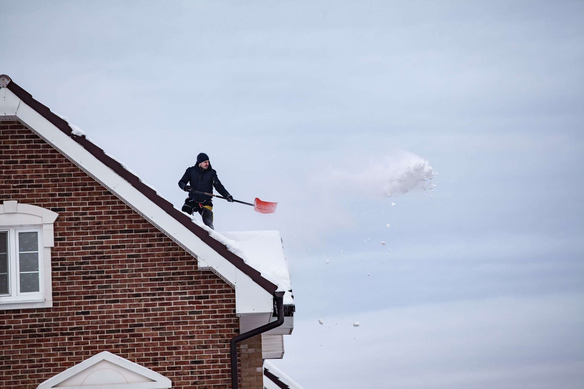 Person shoveling snow from a snowy roof beside a brick house