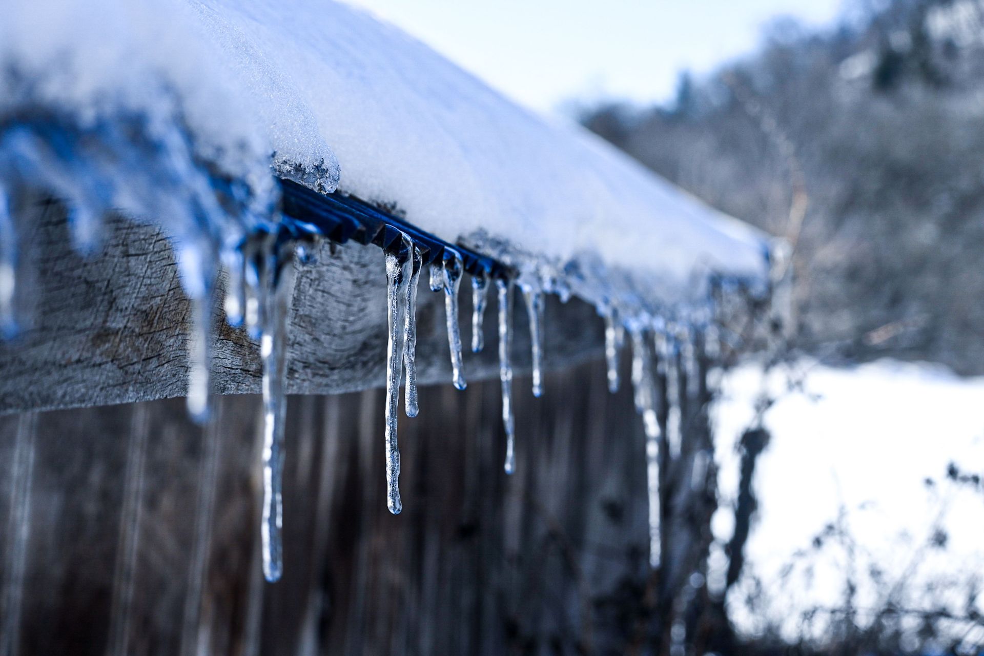 Icicles hanging from a snow-covered roof edge beside a blurred winter landscape